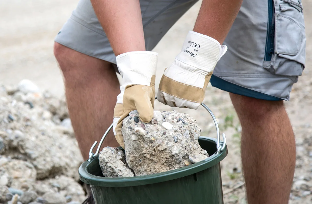 Behandschuhte Hände tragen den olivgrünen Stall- und Baueimer mit Bauschutt