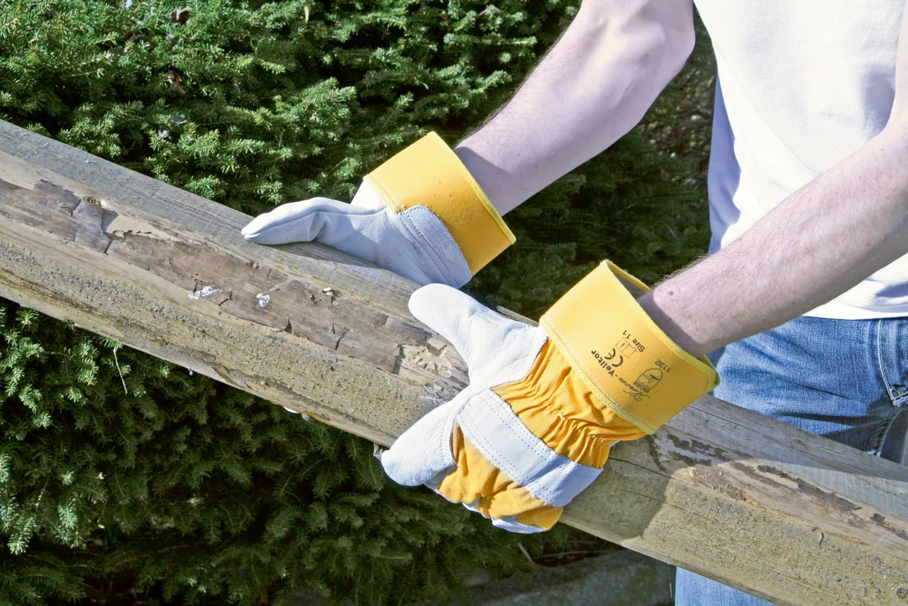 Behandschuhte Hände halten einen Holzbalken, Hecke im Hintergrund