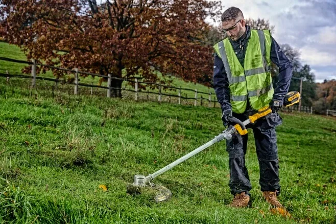 Mann trimmt mit dem DeWalt DCMST561N Akku-Rasentrimmer eine Wiese (Warnweste)