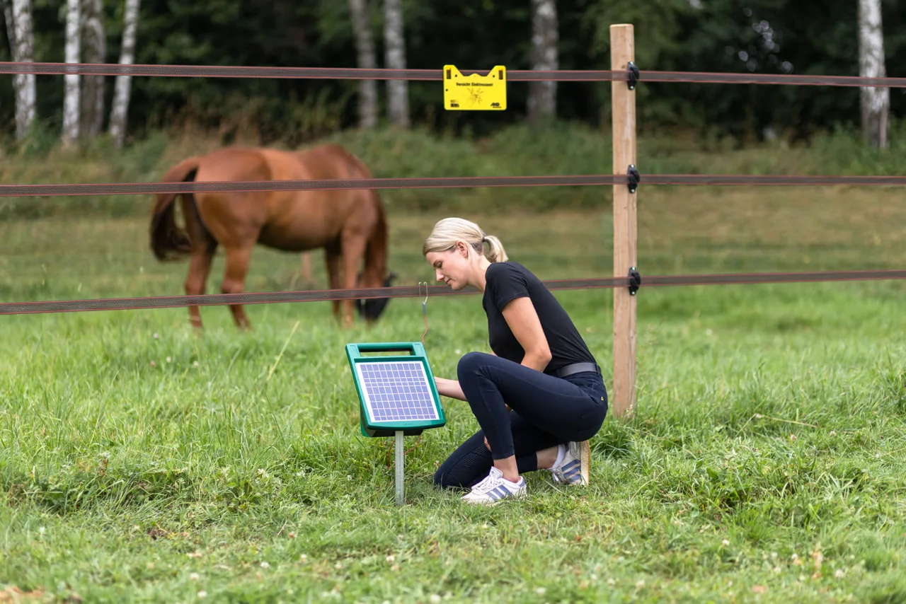 Frau kniet am SunPower-Solargerät am Pfosten, Pferd dahinter