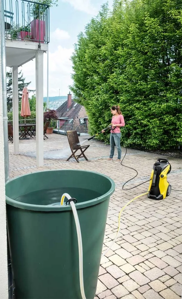 Kärcher-Hochdruckreiniger saugt über den SH 5 Schlauch Wasser aus einer Regentonne, Frau reinigt die Terrasse