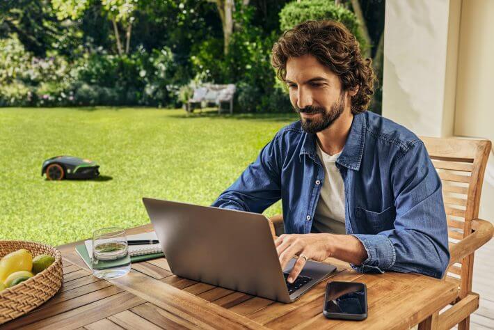 Mann am Laptop im Garten, STIHL iMOW 7 EVO Mähroboter im Hintergrund