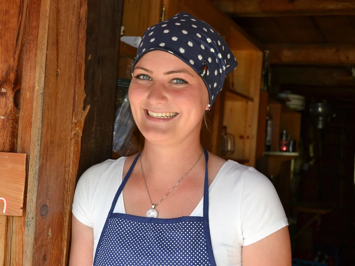 Frau mit blauem Hairtex Stall-Beanie (Edelweiß-Muster) und Schürze vor einer Almhütte