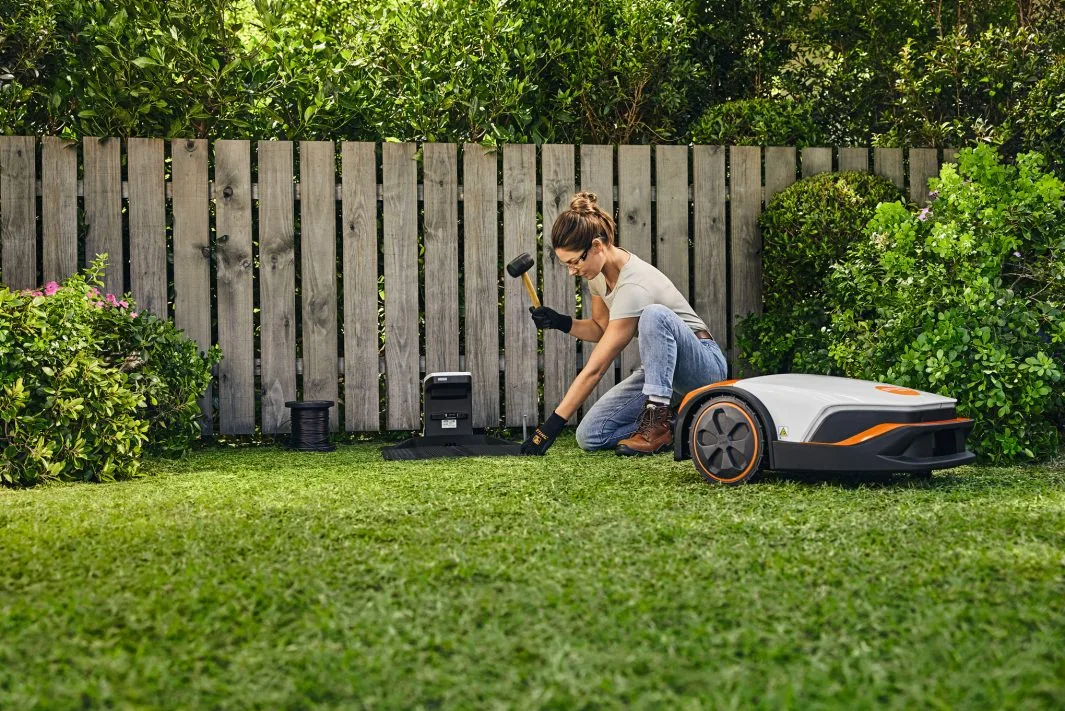 Frau verlegt den STIHL iMOW Begrenzungsdraht im Garten neben dem Mähroboter